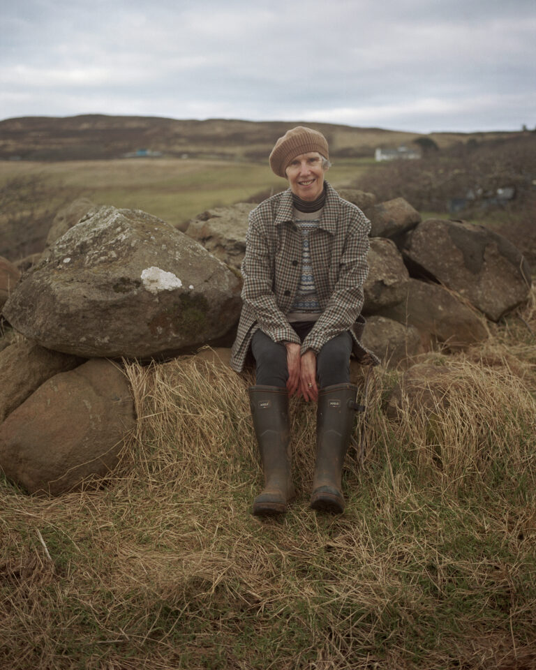 Lucy Montgommery sits amongst a rugged skye landscape. She wears a weaver’s jacket by Johnstons of Elgin, and cashmere Fair Isle jumper & beret, both Brora.